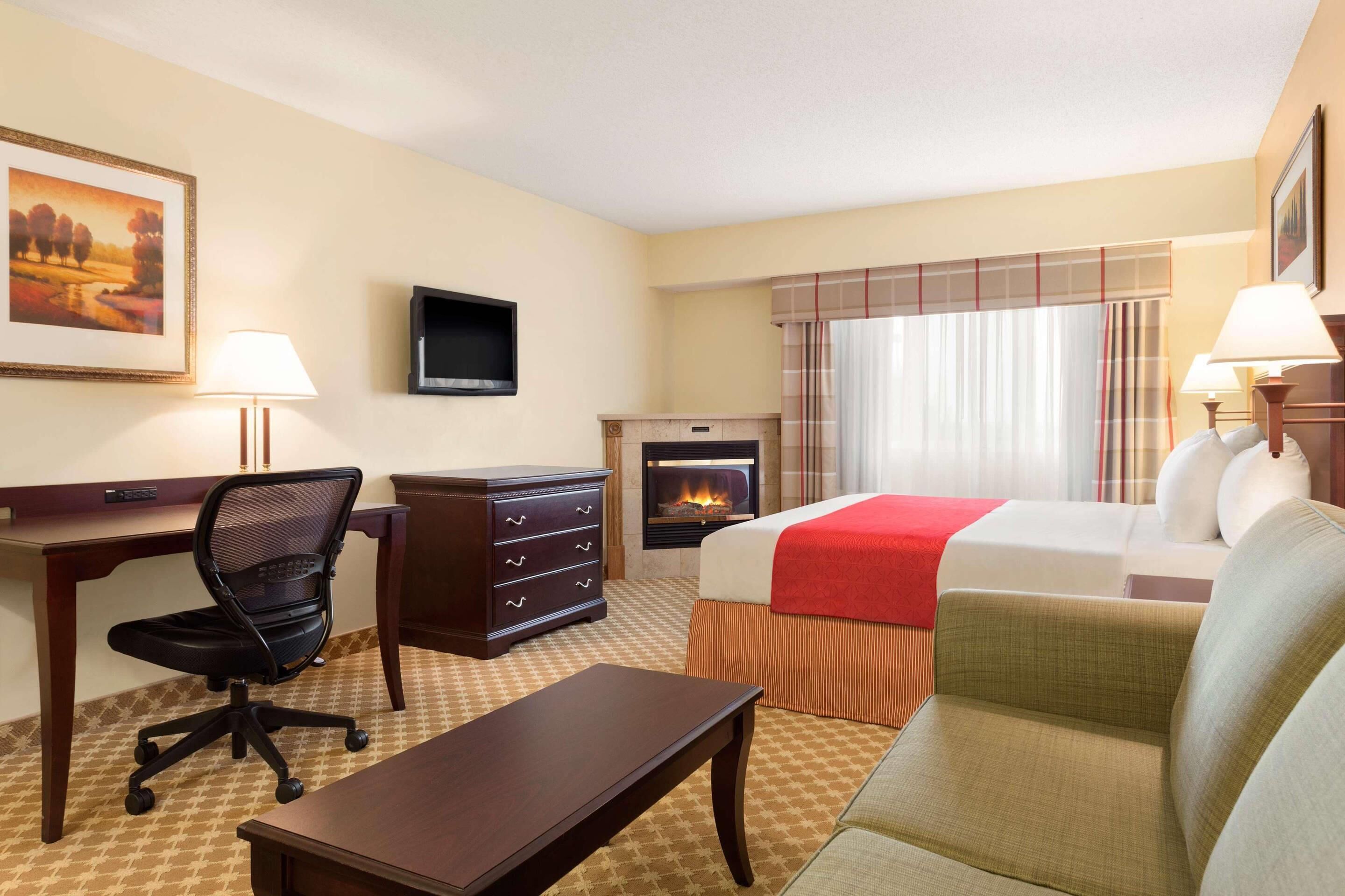 A hotel room with a bed, a desk with a chair, a dresser, a television, and a fireplace.