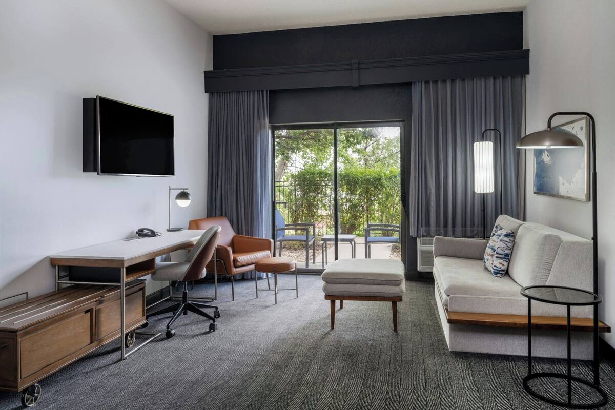 A modern hotel room with a flat-screen TV, a desk with a chair, a sofa, a small ottoman, and a side table.