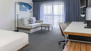 Ein Hotelzimmer mit Bett, Sofa, Schreibtisch, Stuhl, Fernseher und einem Fenster mit Vorhängen.