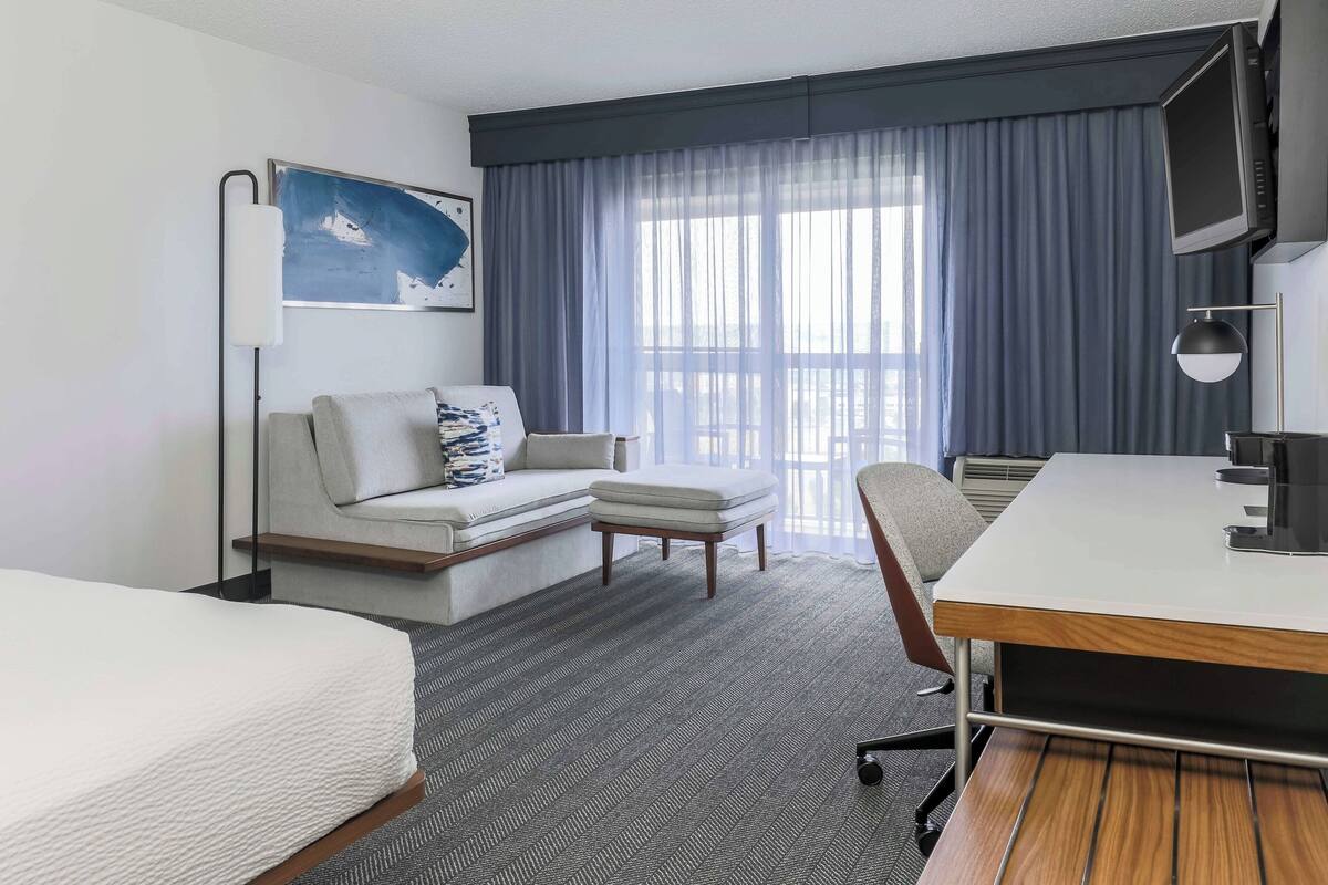 A hotel room with a bed, a sofa, a desk, a chair, a TV, and a window with curtains.