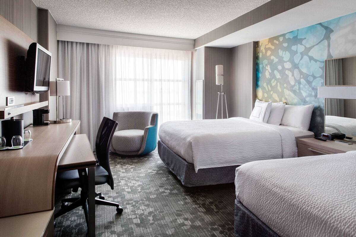 Een hotelkamer met een bureau, twee bedden, een stoel, een televisie en een groot abstract schilderij aan de muur.