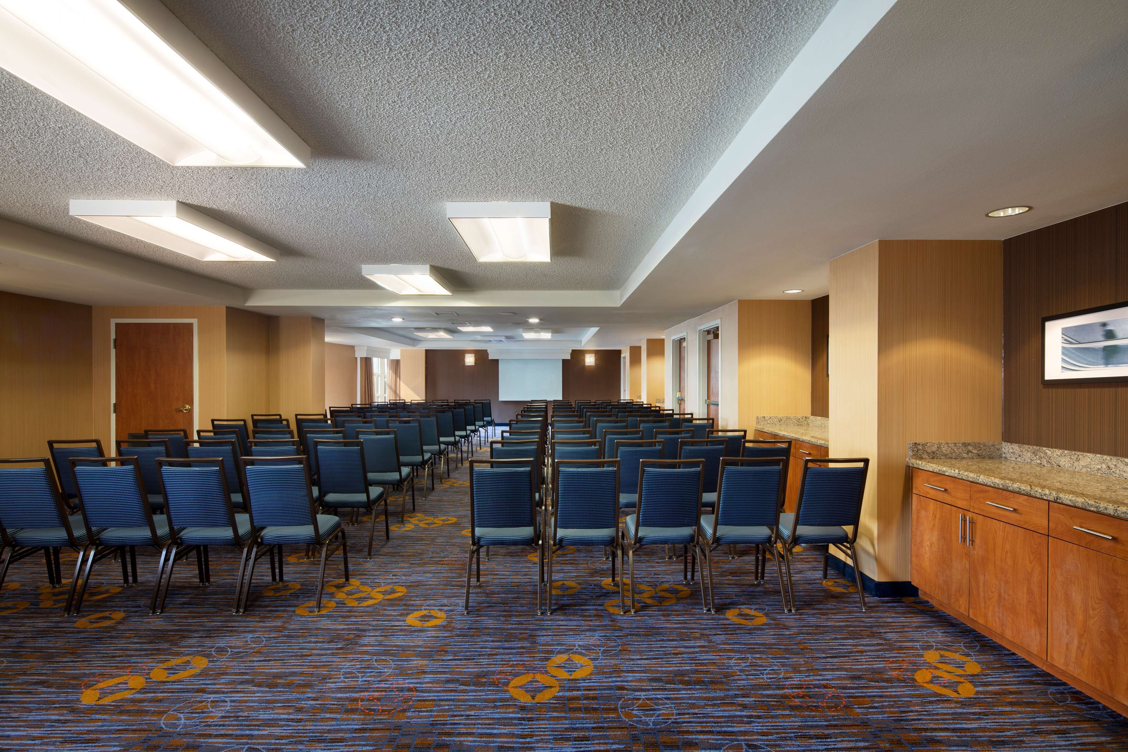 Une salle de conférence avec des rangées de chaises, une moquette à motifs, des murs en bois et un grand écran.