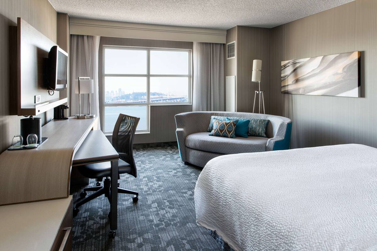 Een hotelkamer met een bed, een bureau met stoel, een televisie en een groot raam met uitzicht over de stad.