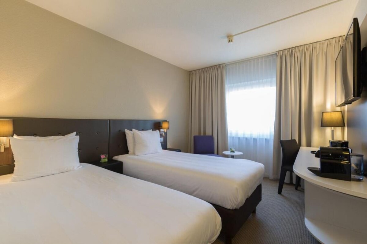Une chambre d’hôtel avec deux lits, un bureau, une chaise, une télévision et une fenêtre avec des rideaux.
