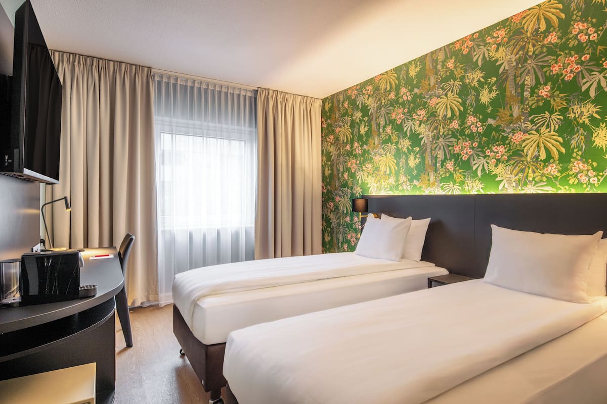 Een hotelkamer met twee bedden, een bureau met computer, een televisie en een groot raam met gordijnen.
