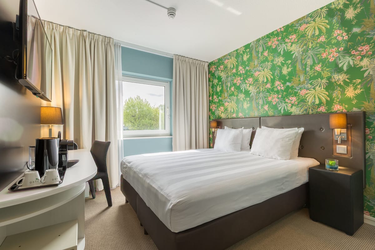 Una habitación de hotel con cama, escritorio, silla, televisión y ventana con cortinas.