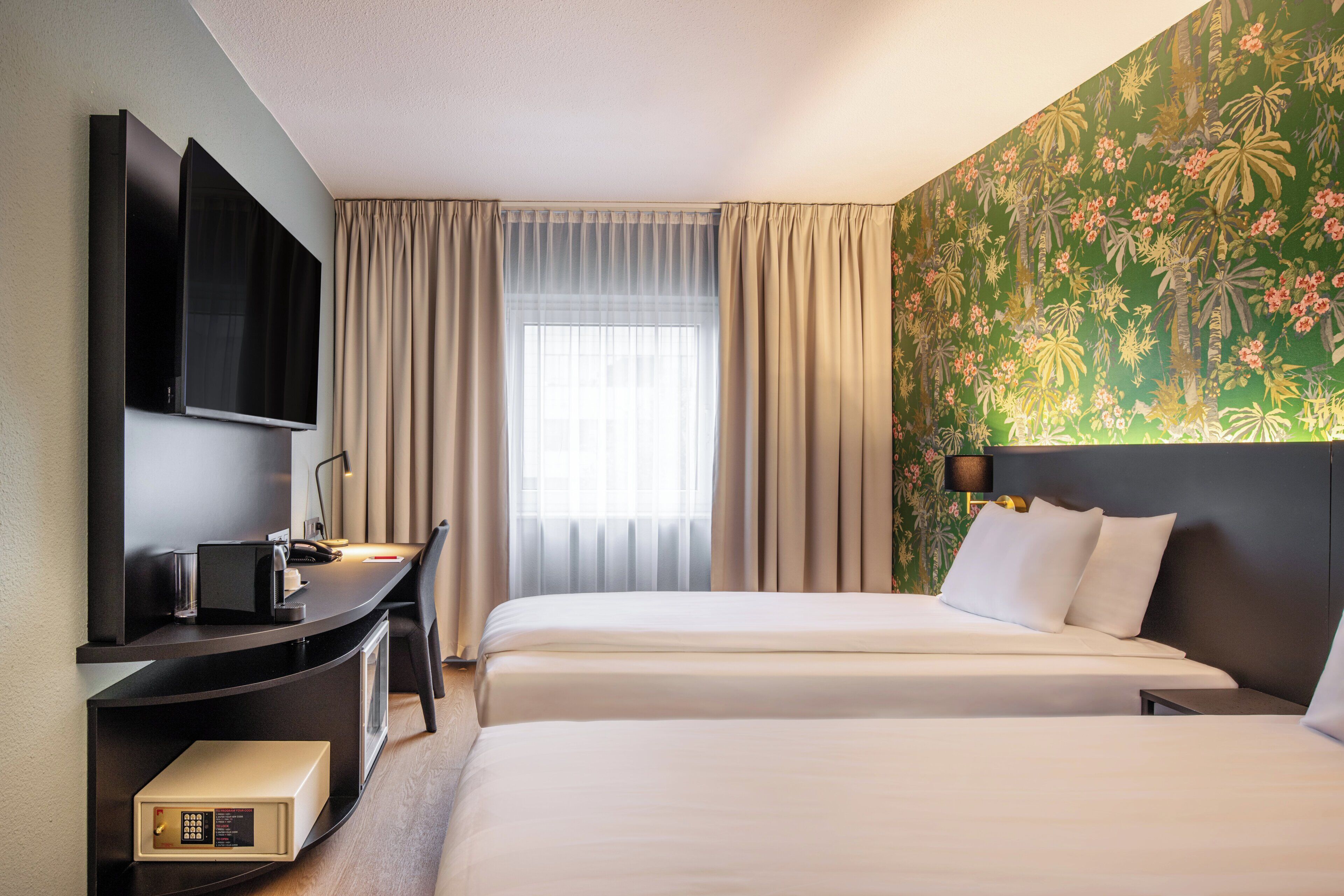 A hotel room with two beds, a desk with a flat-screen TV, a minibar, and a large window with curtains.