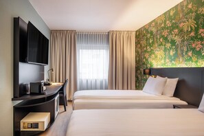 Ein Hotelzimmer mit zwei Betten, einem Schreibtisch, einem Flachbildfernseher, einer Minibar und einem großen Fenster mit Vorhängen.
