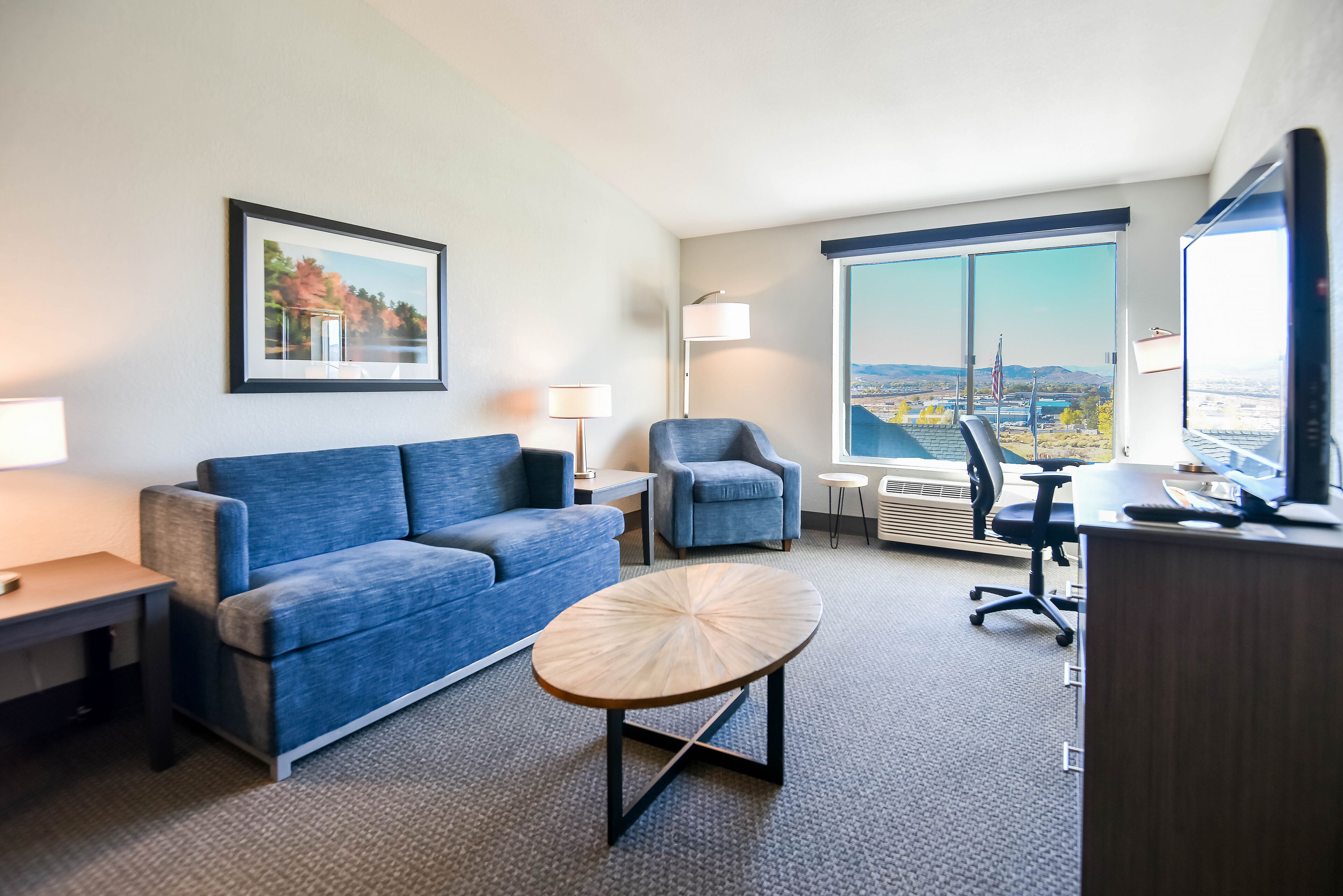 A modern hotel room with a blue sofa, a round coffee table, a desk, a chair, and a large window offering a city view.