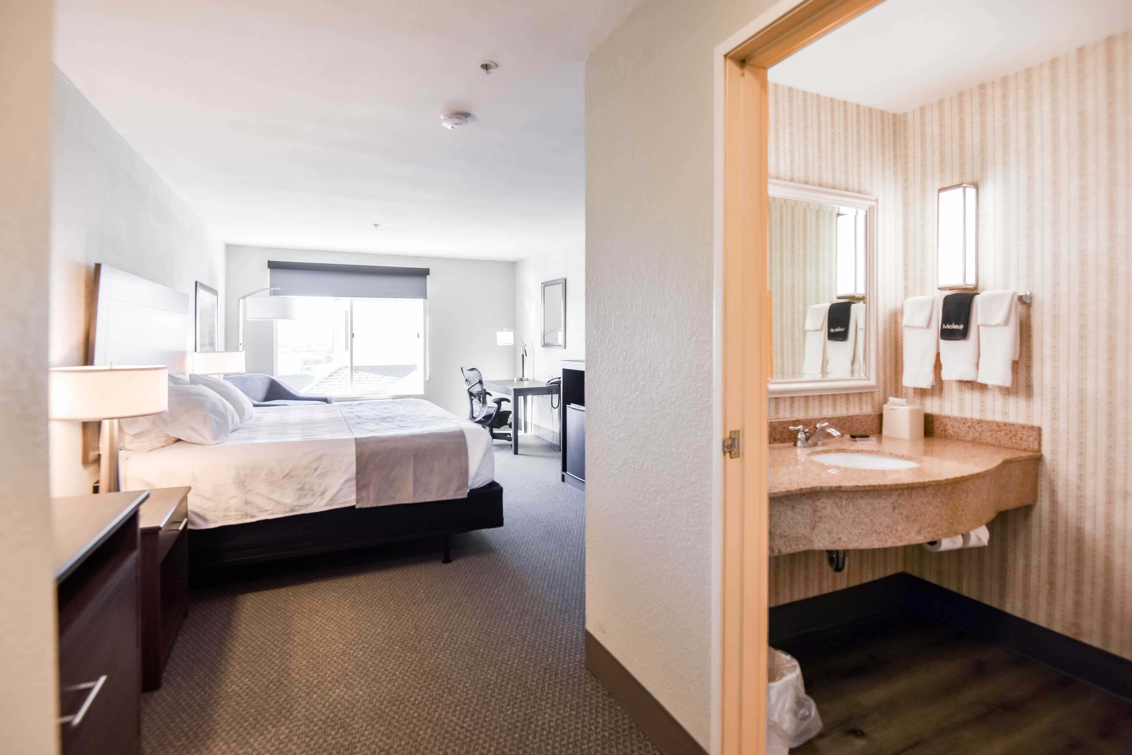 A hotel room with a bed, bedside table, lamp, and a view of the hallway.