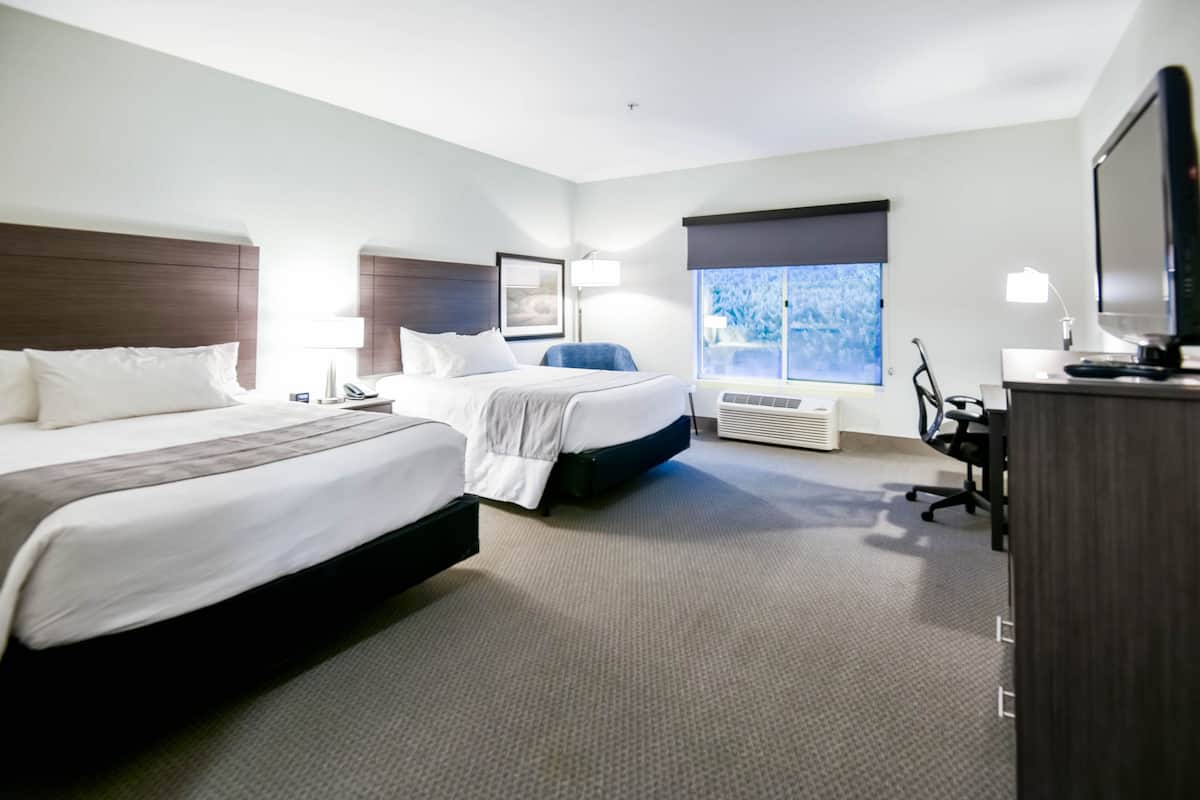 A hotel room with two beds, a desk with a computer, a TV, and a large window with a view of trees.