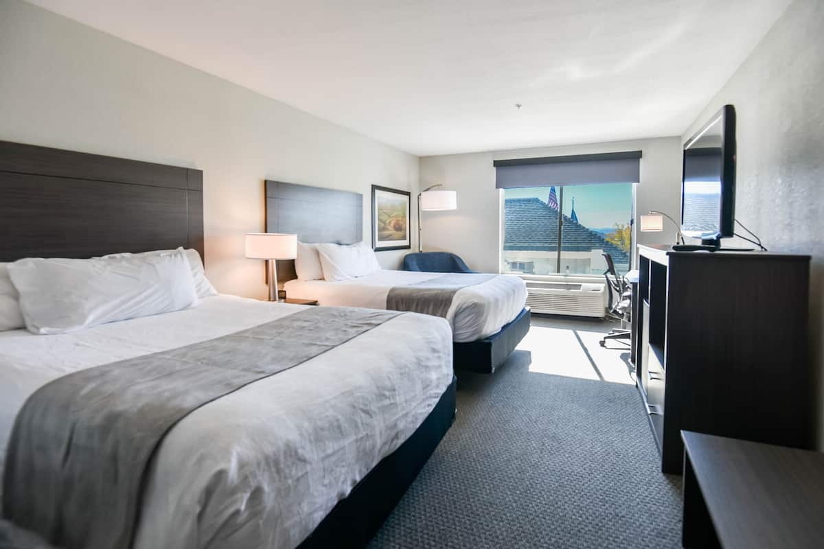 A hotel room with two beds, a desk, a TV, and a large window with a view of a building.