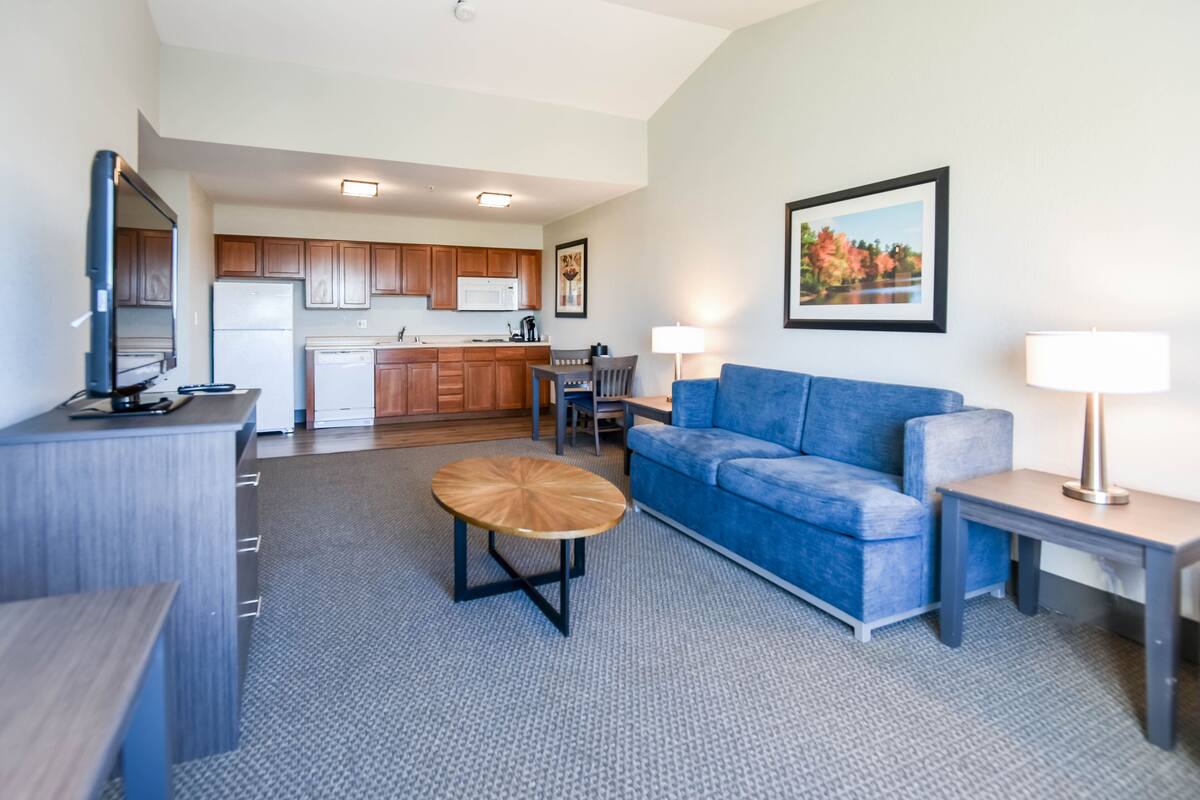A modern living room with a blue sofa, a coffee table, and a TV on a stand. There is a kitchen area in the background with wooden cabinets and a refrigerator.