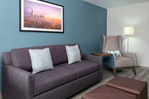 A room with a purple sofa, a chair, a small table, and a framed cityscape picture on the wall. - Hilton Garden Inn Charlotte Uptown (Charlotte)