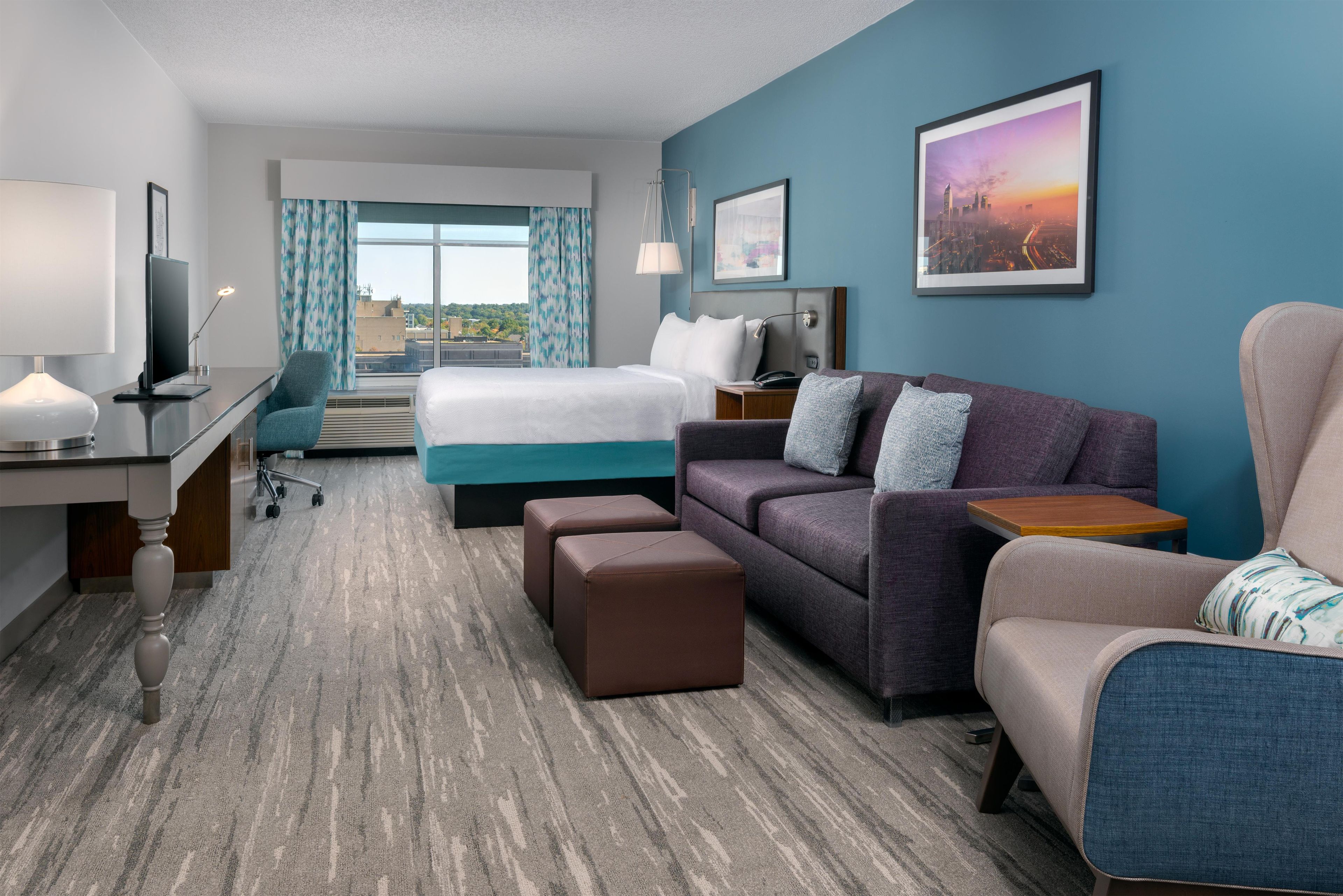 A hotel room with a bed, a sofa, a desk, a chair, and a view of the city.