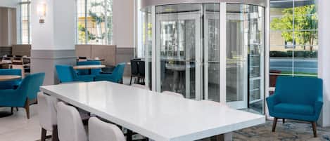 Un hall d’hôtel moderne avec une table blanche centrale entourée de chaises, une cloison en verre et de grandes fenêtres offrant une vue sur l’extérieur.