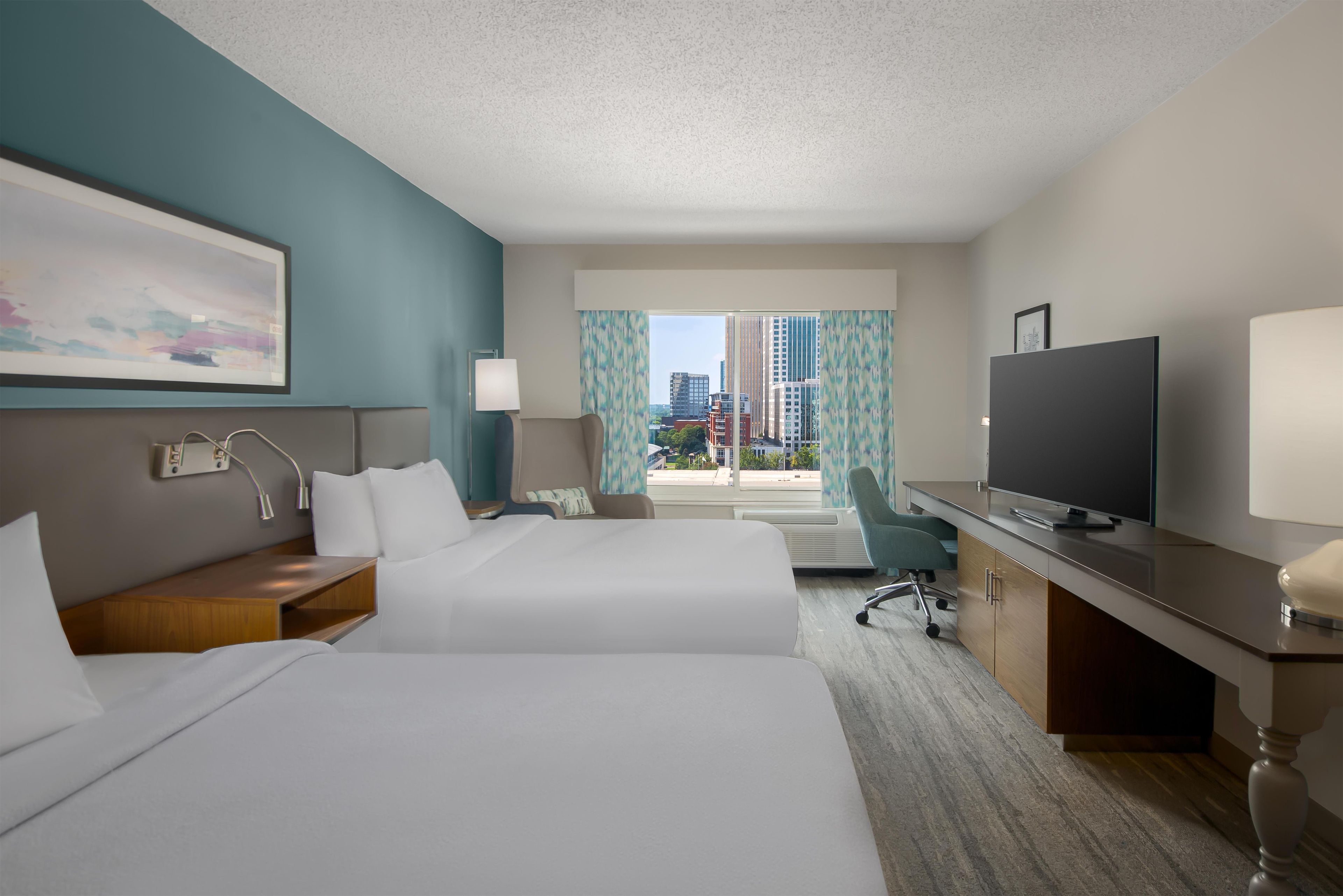 Ein Hotelzimmer mit zwei Betten, einem Schreibtisch, einem Fernseher und Blick auf die Stadt.