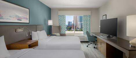 Ein Hotelzimmer mit zwei Betten, einem Schreibtisch, einem Fernseher und Blick auf die Stadt.