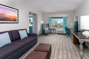 A hotel room with a sofa, a small table, a chair, a desk with a lamp, and a large window with curtains. - Hilton Garden Inn Charlotte Uptown (Charlotte)
