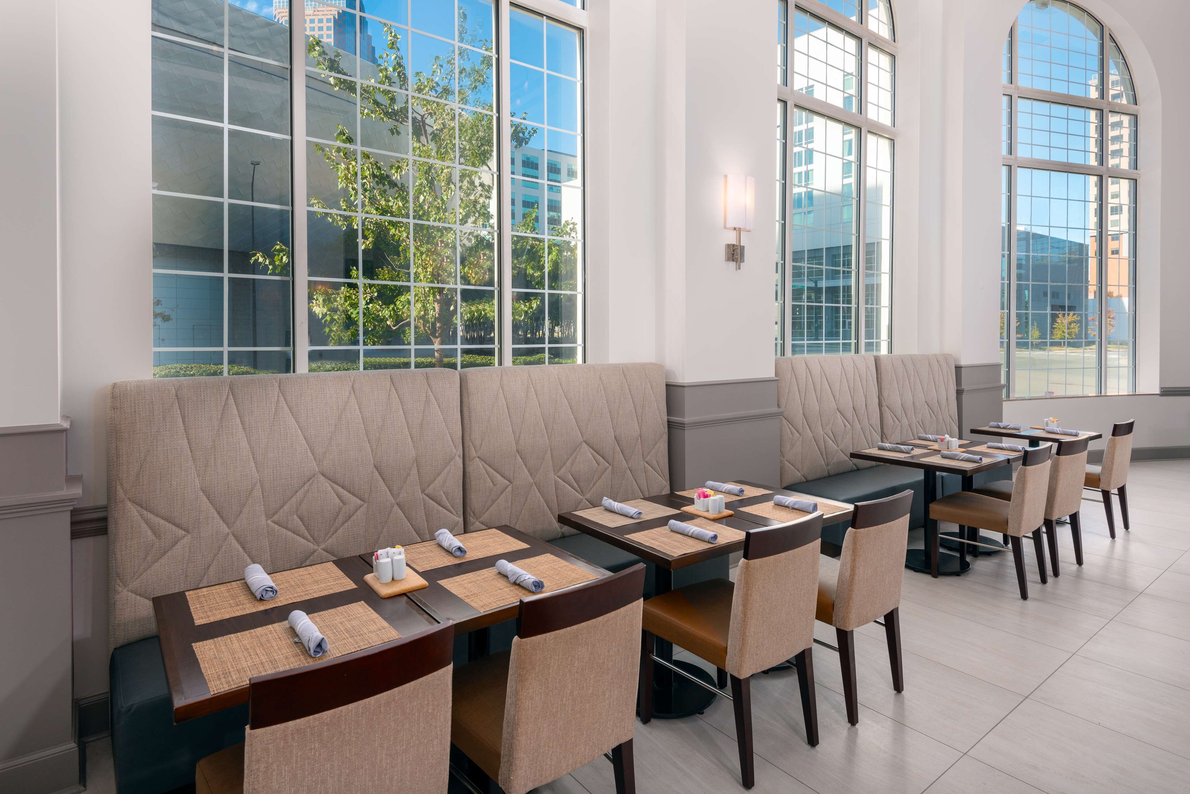 Un comedor moderno con ventanales altos, asientos acolchados y mesas ordenadas con manteles individuales y cubiertos.