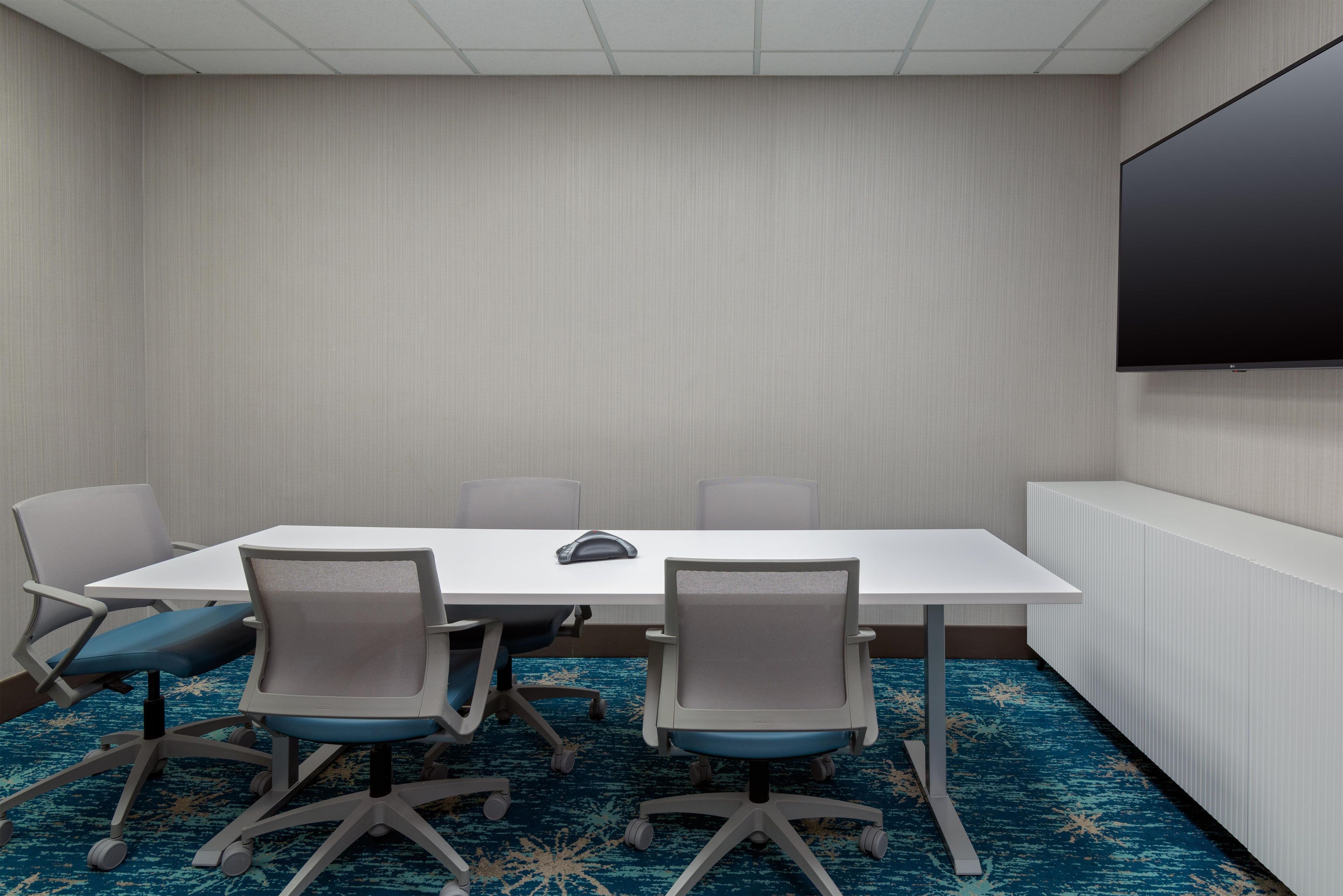 A conference room with a white table, four chairs, a flat-screen TV on the wall, and a patterned carpet.