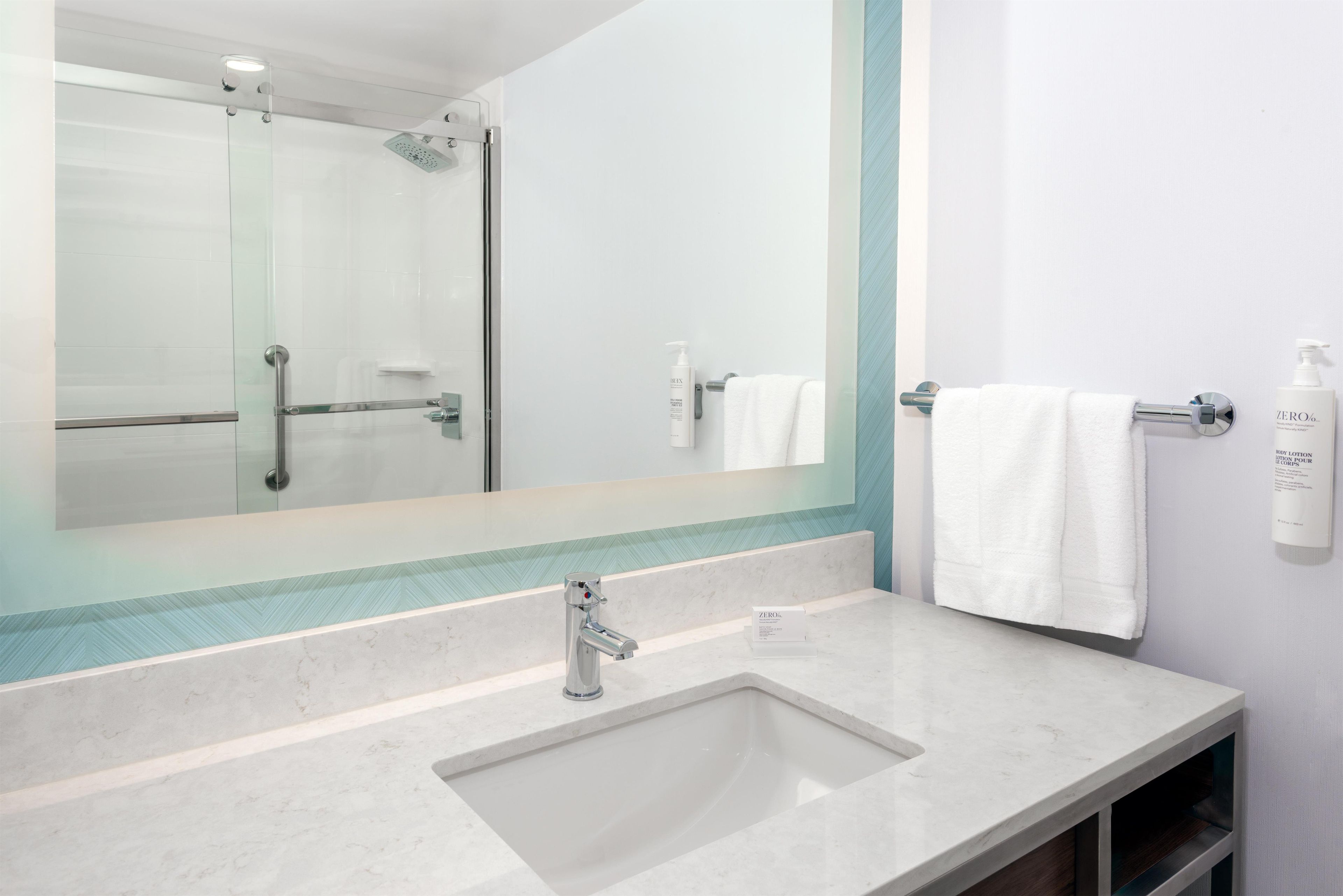 Ein Badezimmer mit Duschkabine aus Glas, einem Waschbecken mit Chromarmatur und einem Spiegel, der die Dusche reflektiert.
