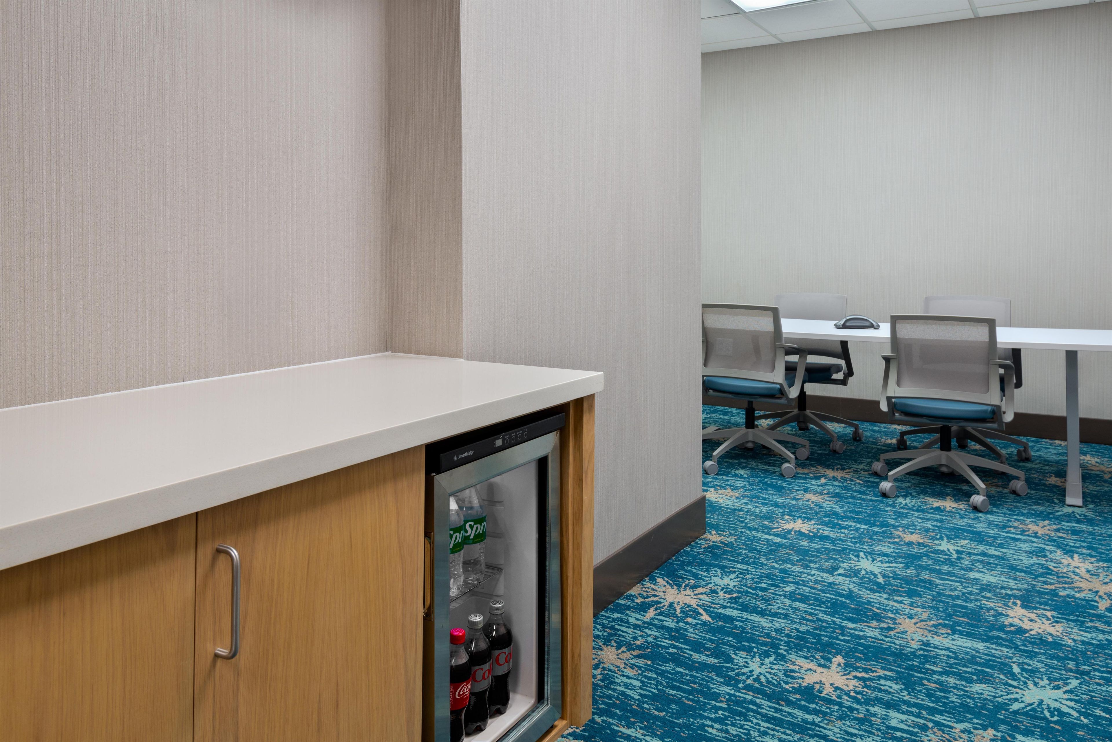 A conference room with a white table, four chairs, and a refrigerator with beverages.