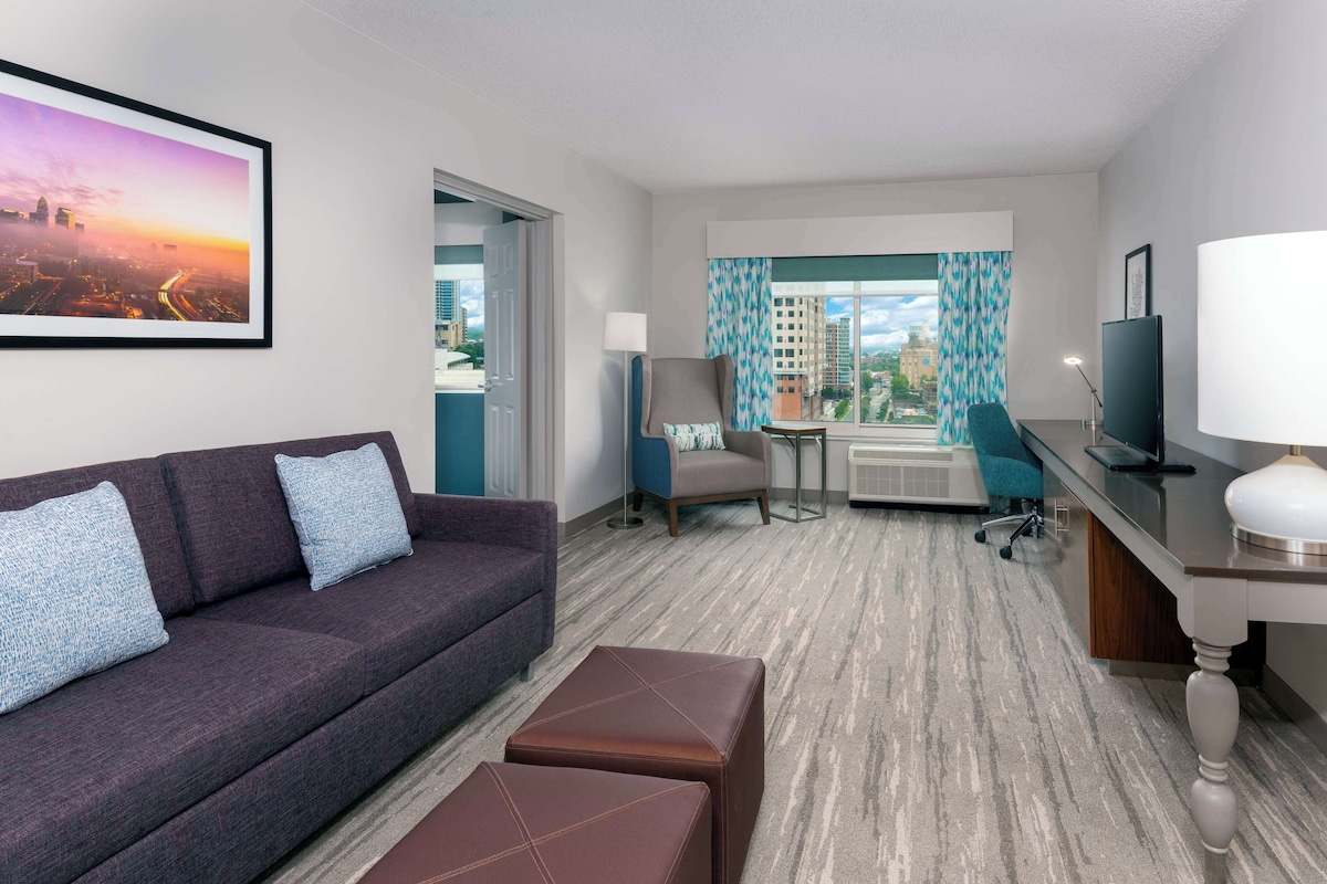 A hotel room with a cityscape view, a sofa, a desk with a lamp, and a chair.