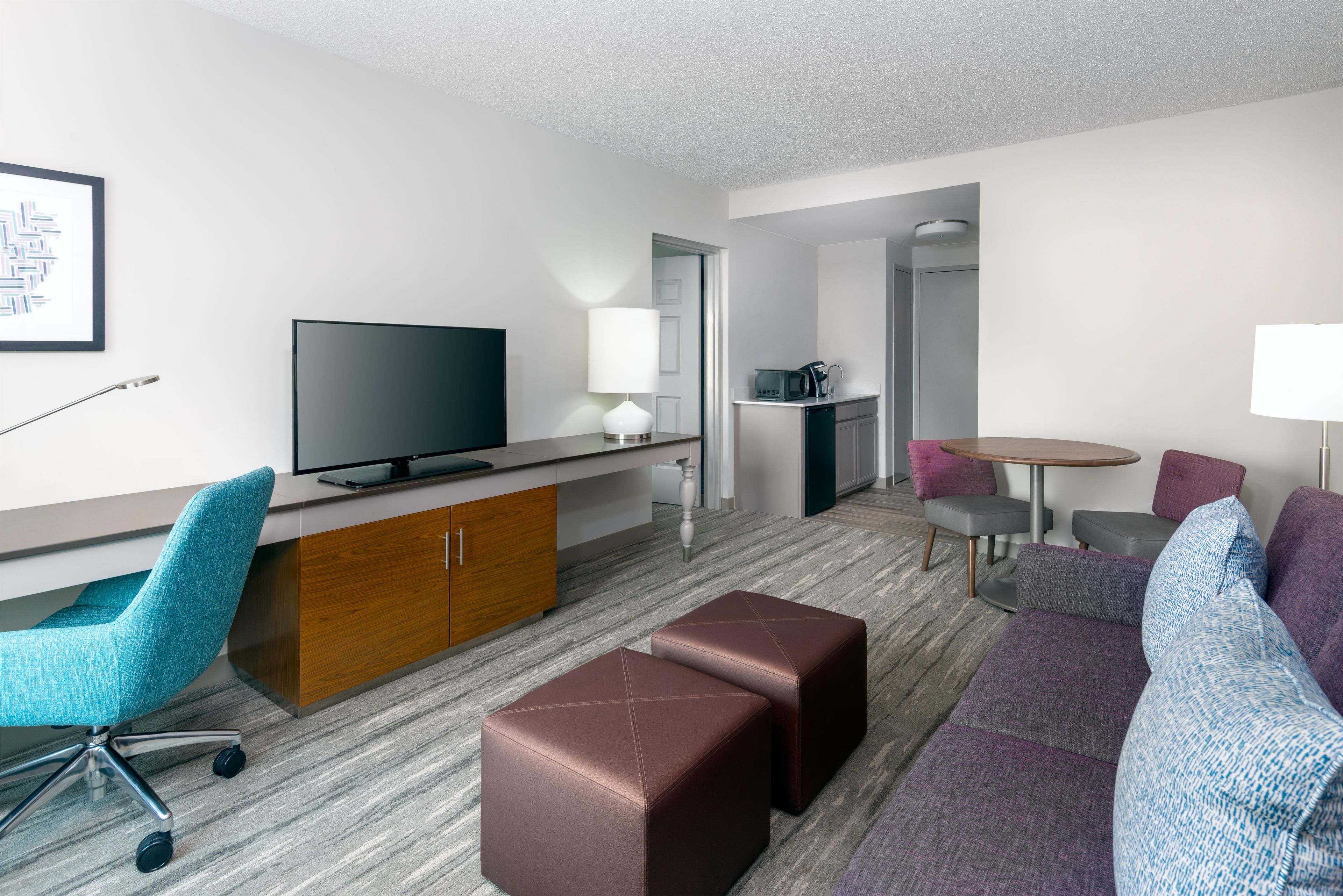 A modern hotel room with a flat-screen TV, a desk, a sofa, a coffee table, and a small kitchenette.