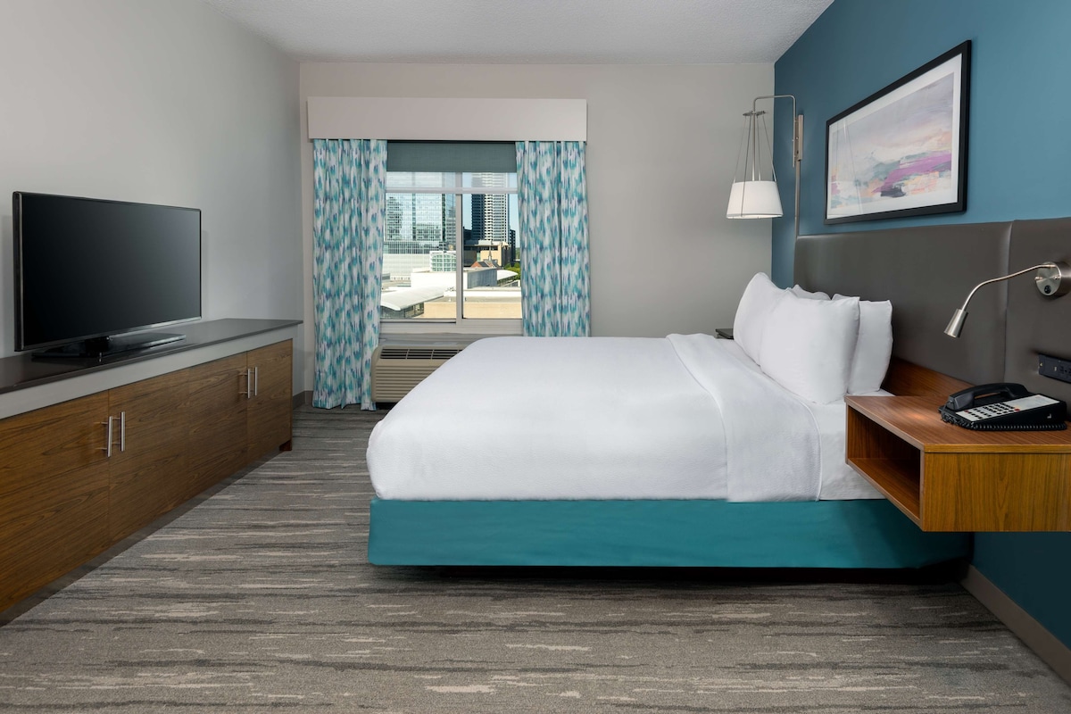 A modern hotel room with a large bed, a flat-screen TV, a desk with a lamp, and a city view through the window.