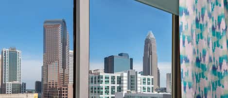 Un paisaje urbano a través de una ventana, con edificios modernos, cielo despejado y cortina estampada.
