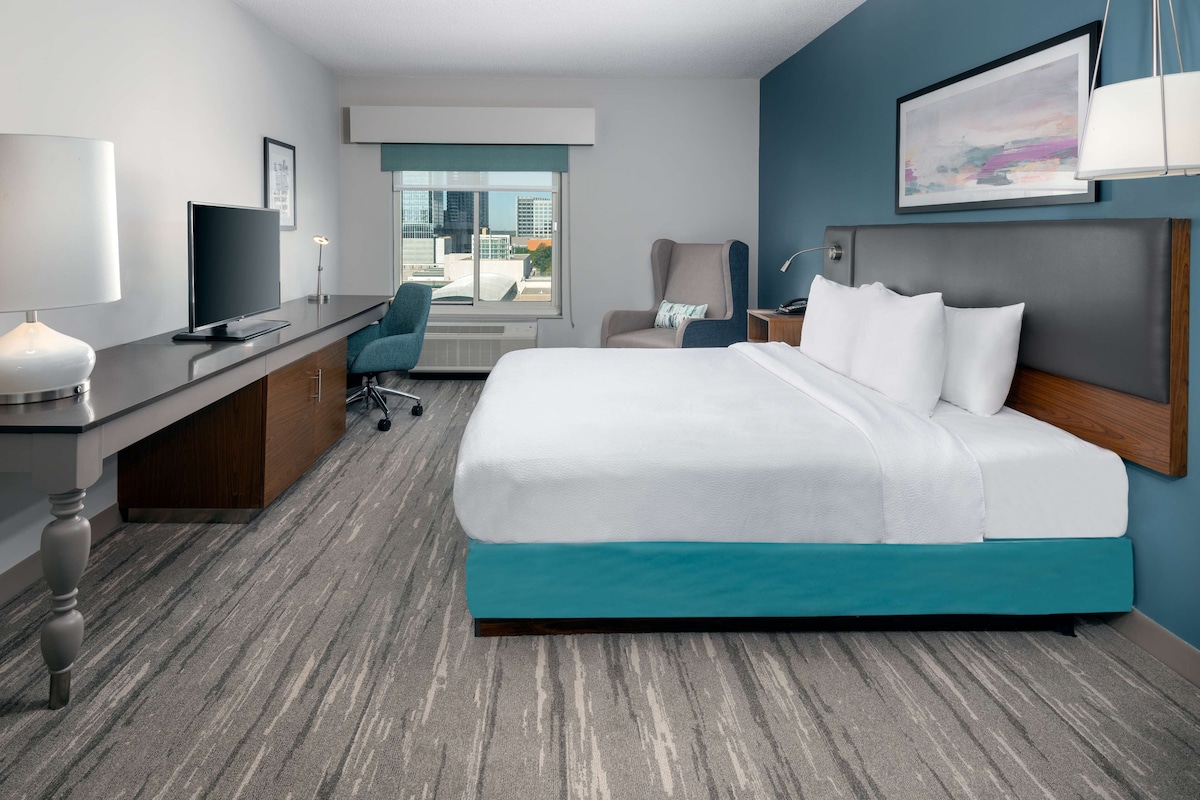 A hotel room with a bed, desk, chair, and a view of the city through the window.