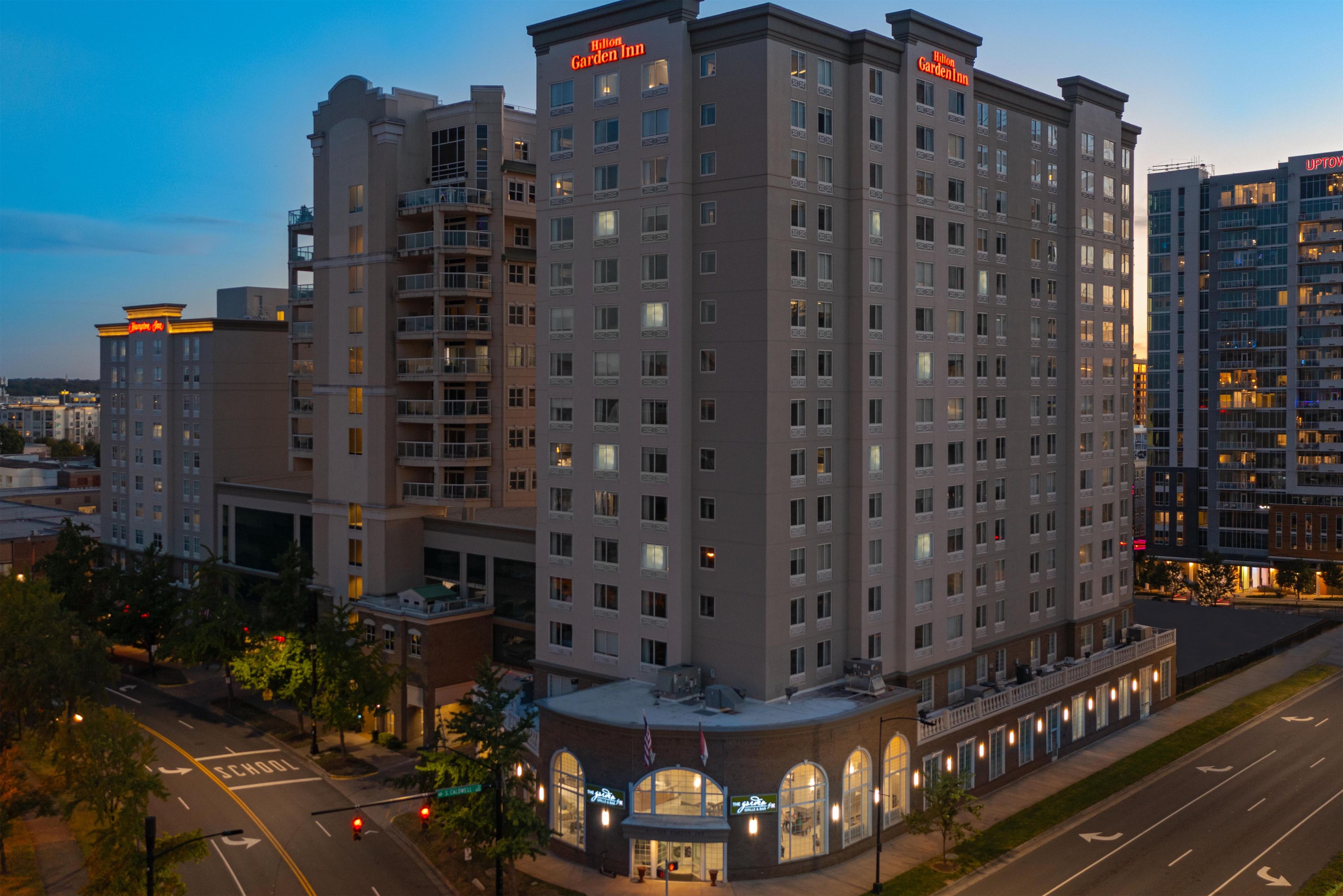 Un hotel de varios pisos con un letrero visible de 'Hilton Garden Inn', ubicación en esquina con vista a la calle y un estacionamiento al frente.