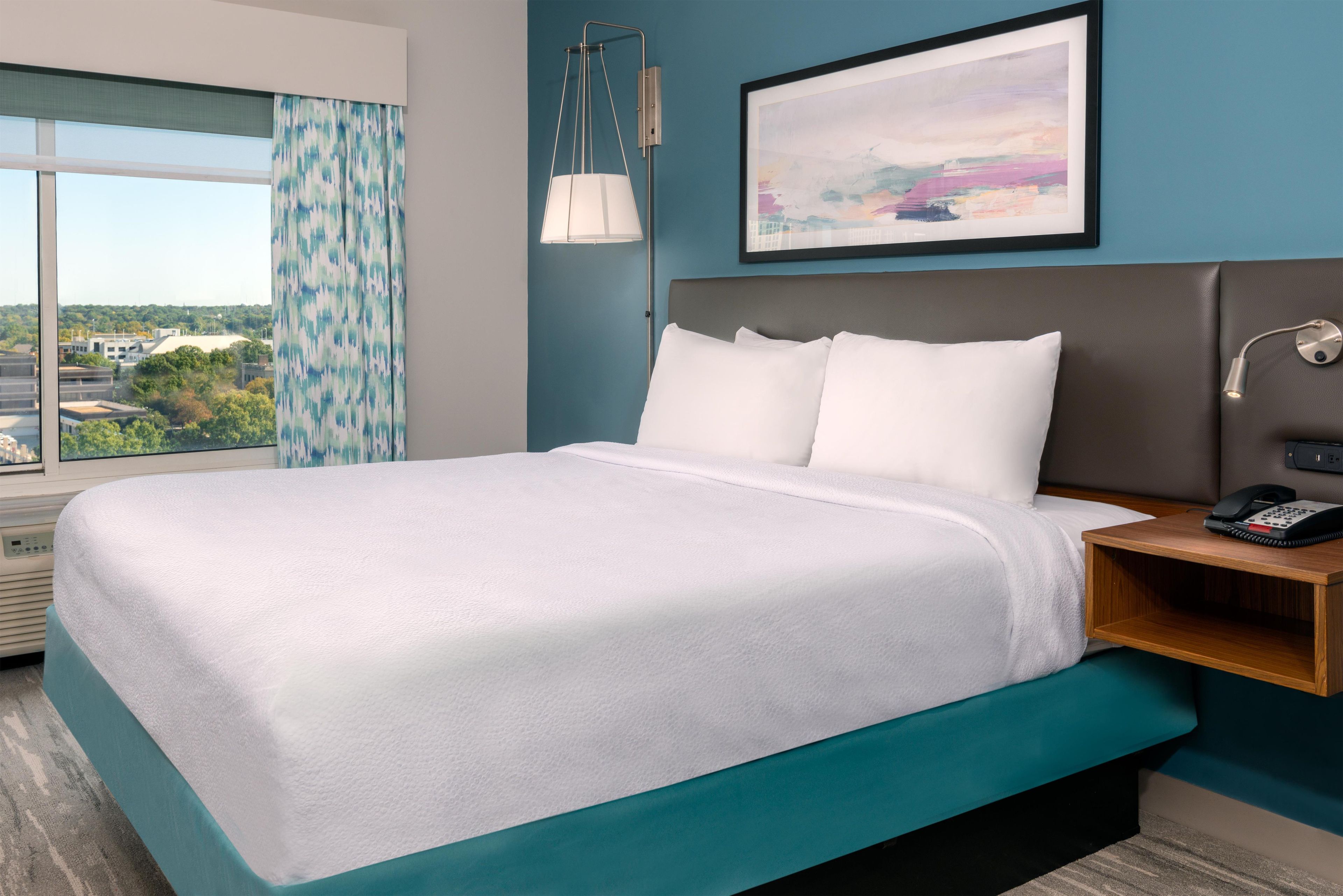 A hotel room with a large bed, a bedside table with a phone, and a view of the city through the window.