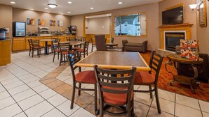A hotel dining area with a large table, multiple chairs, a sofa, a fireplace, a television, and a kitchen area with appliances.