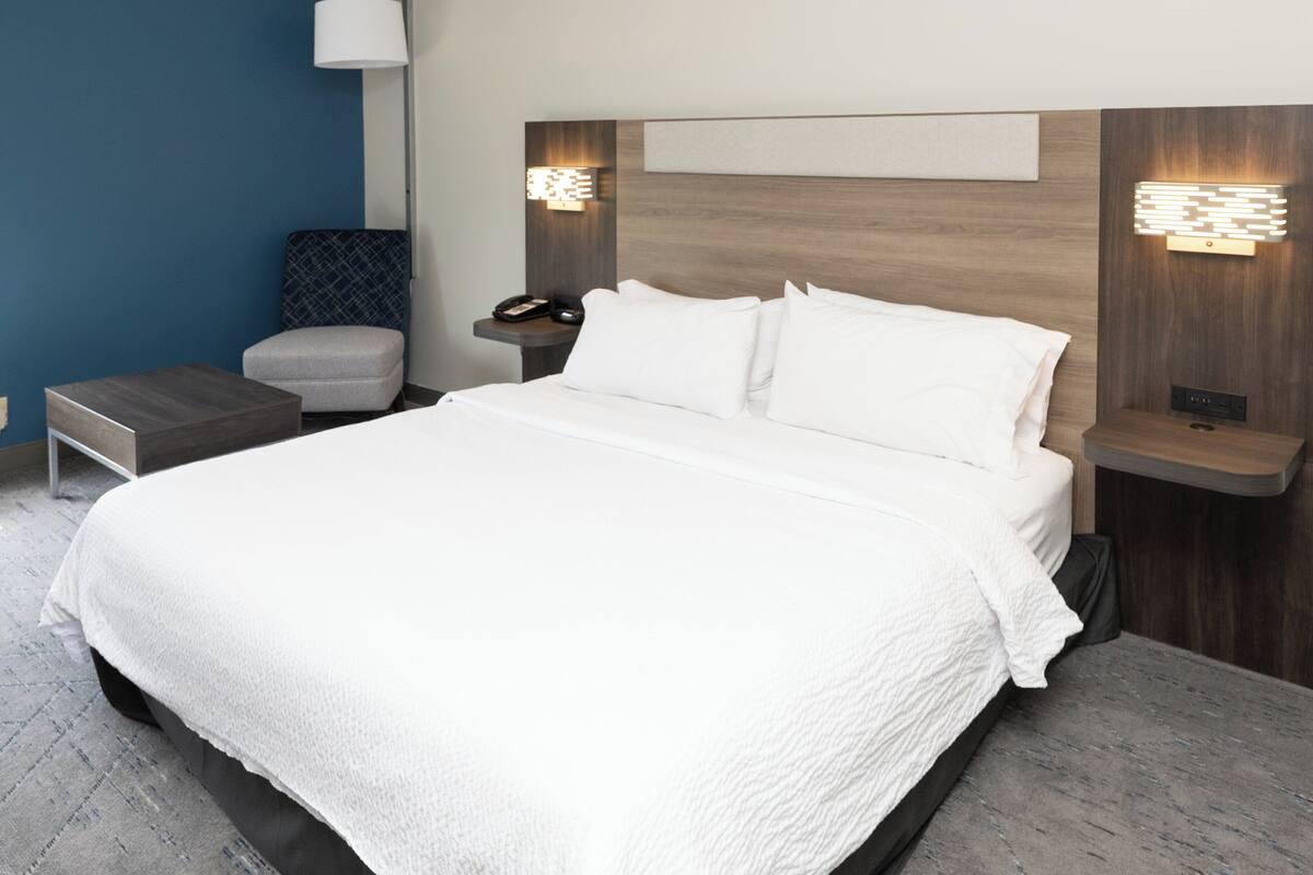 A modern hotel room with a large bed, a nightstand, a chair, and a wall-mounted lamp.