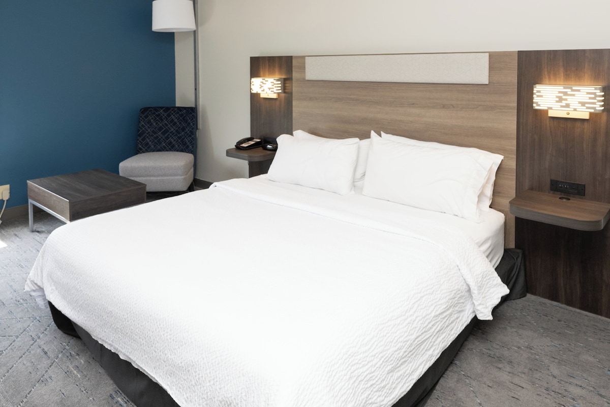 A modern hotel room with a large bed, a nightstand, a chair, and a wall-mounted lamp.