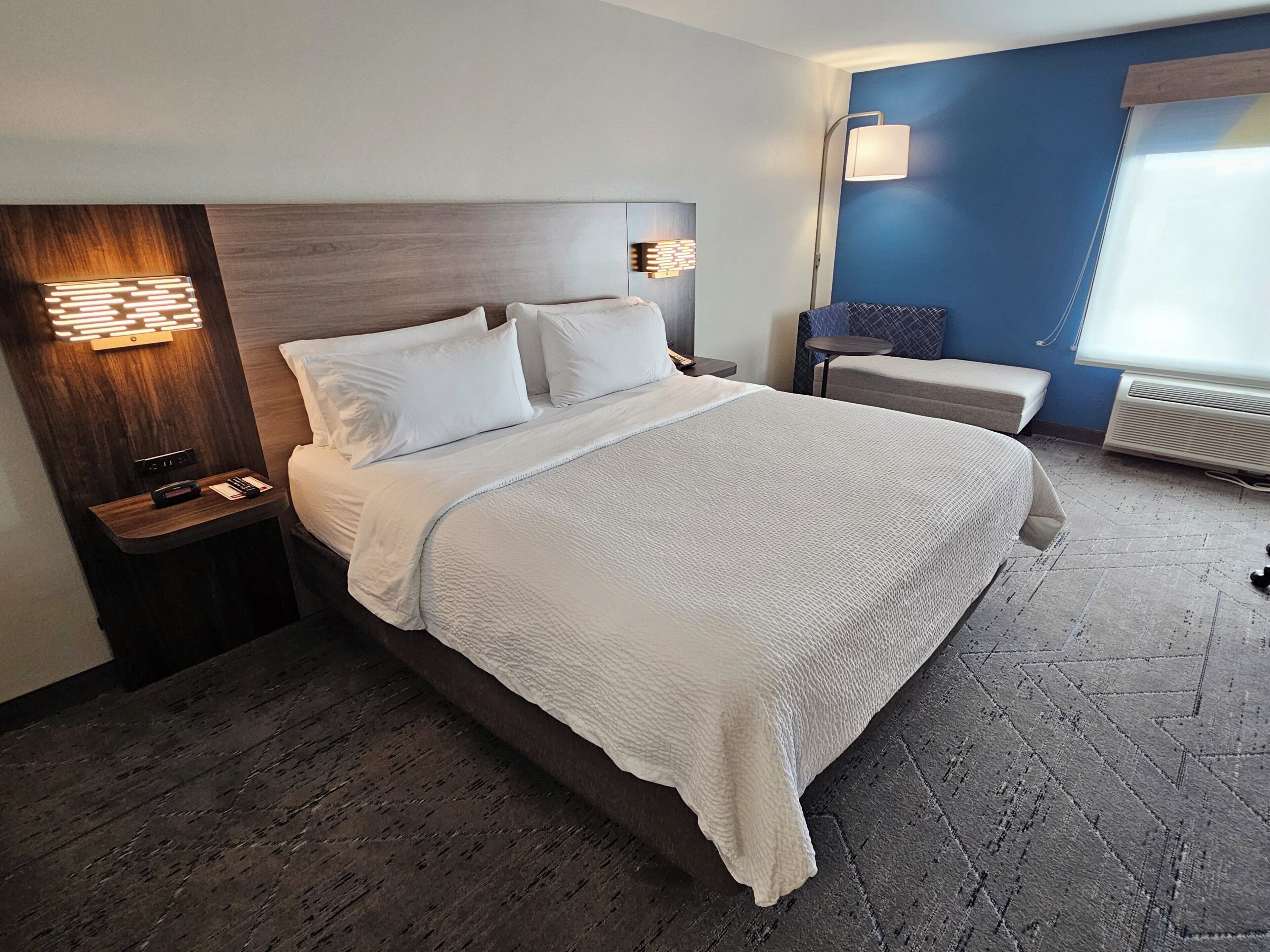 A hotel room with a large bed, a bedside table with a lamp, a small table, and a bench.