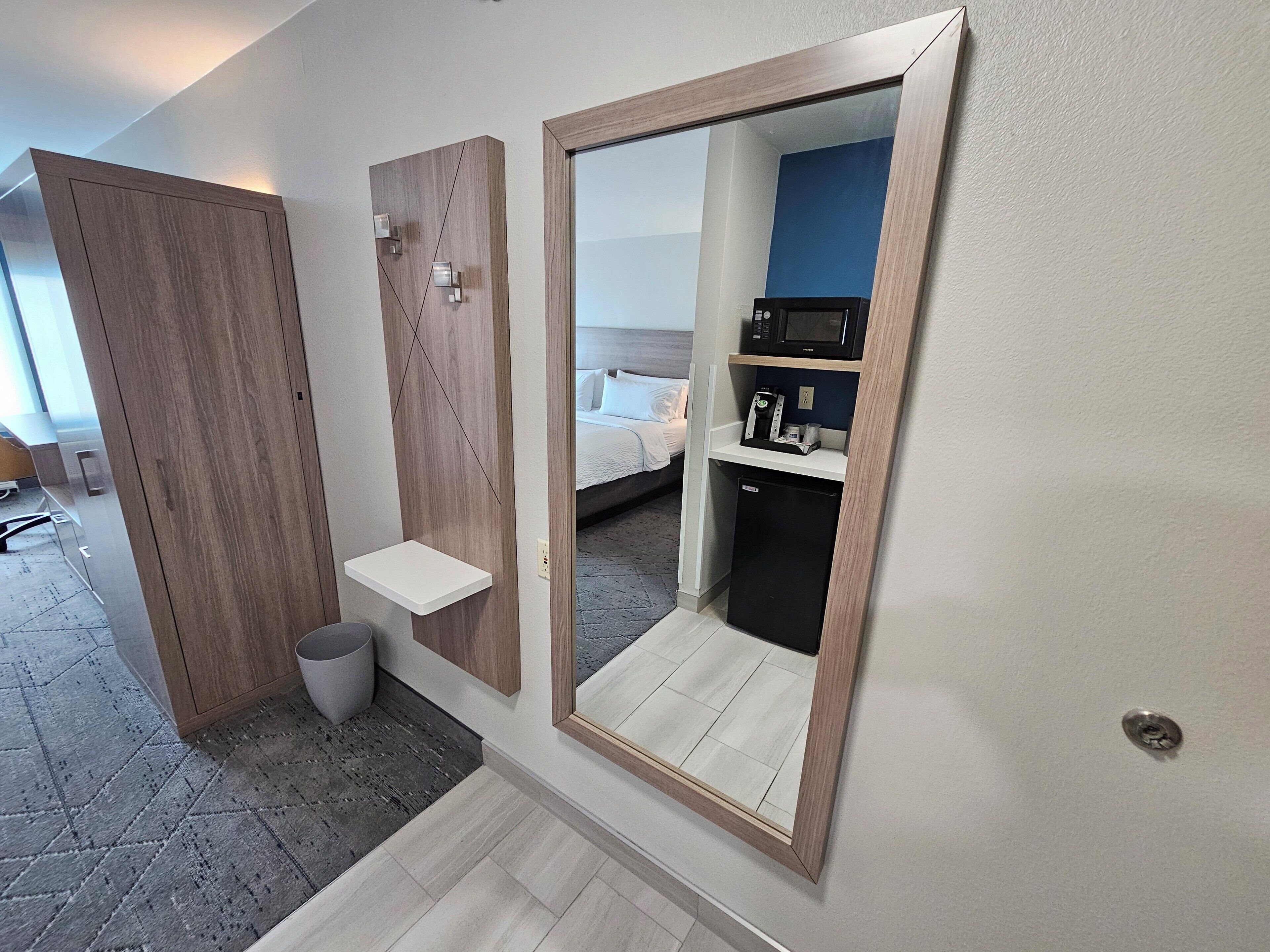 A hotel room with a wooden wardrobe, a small white shelf, a large mirror, and a compact kitchenette with a microwave and coffee machine.