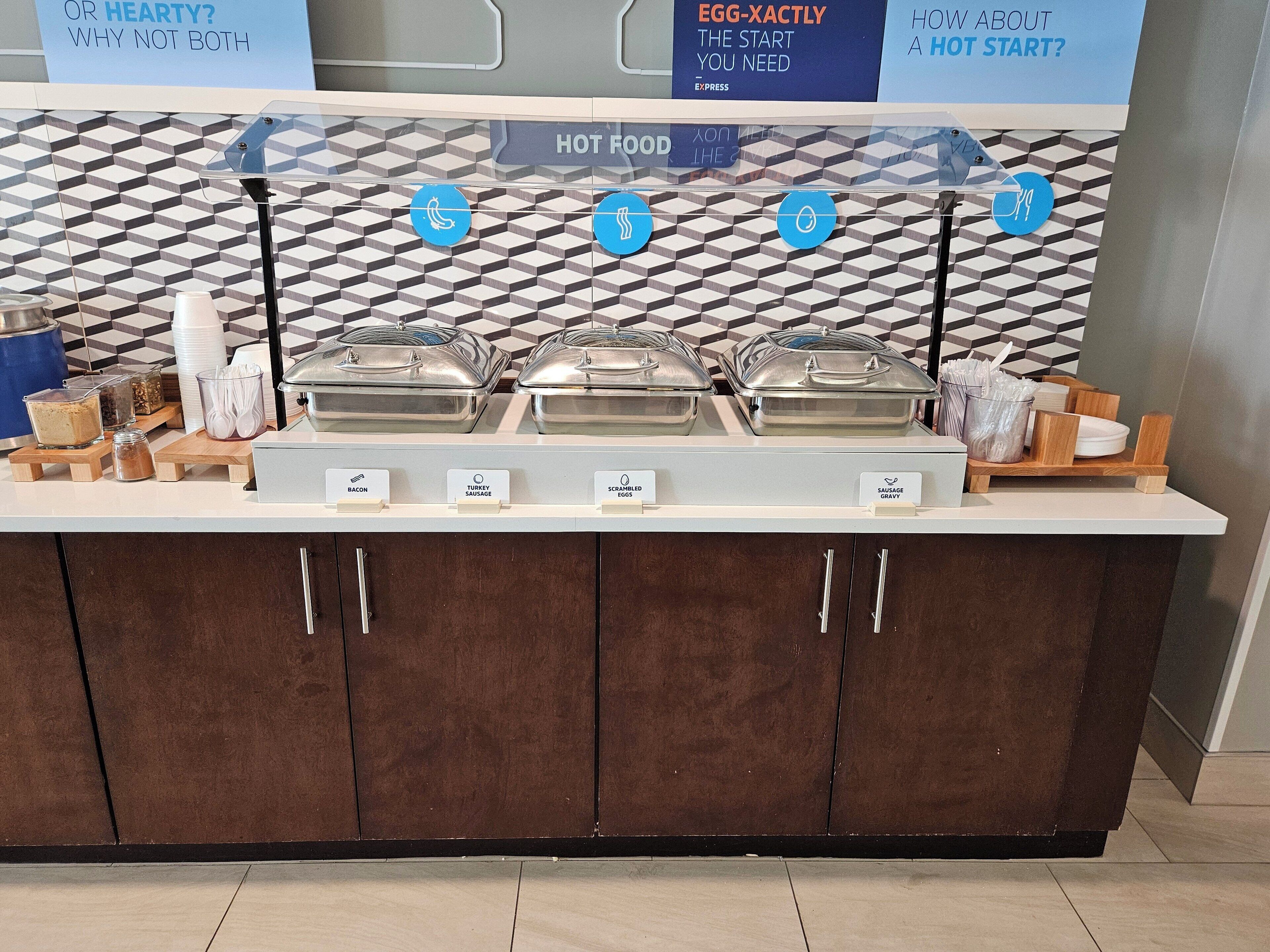 A buffet counter with labeled hot food options including bacon, turkey sausage, scrambled eggs, and sausage gravy.