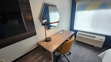 A hotel room with a wooden desk, a chair, a large mirror, a flat-screen TV, and a window with blinds.
