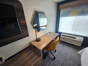 A hotel room with a wooden desk, a chair, a large mirror, a flat-screen TV, and a window with blinds. - Holiday Inn Express Hotel & Suites Cincinnati-Blue Ash by IHG (Cincinnati)