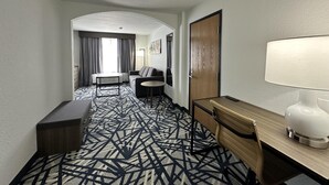 A hotel room with a patterned carpet, a sofa, a desk with a lamp, a chair, and a door.