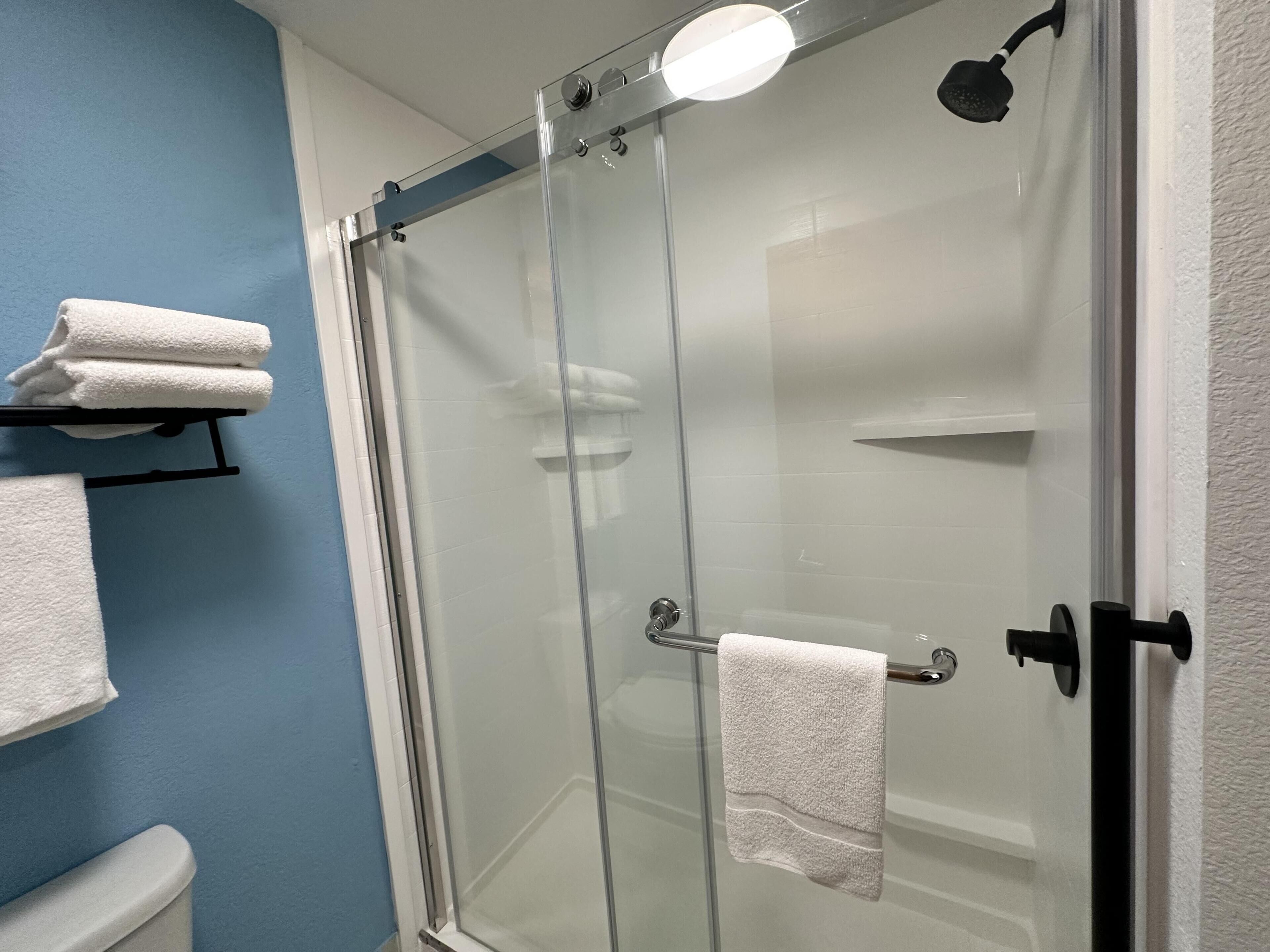 A glass-enclosed shower with a handheld showerhead, a towel rack with folded towels, and a toilet visible in a bathroom with blue walls.