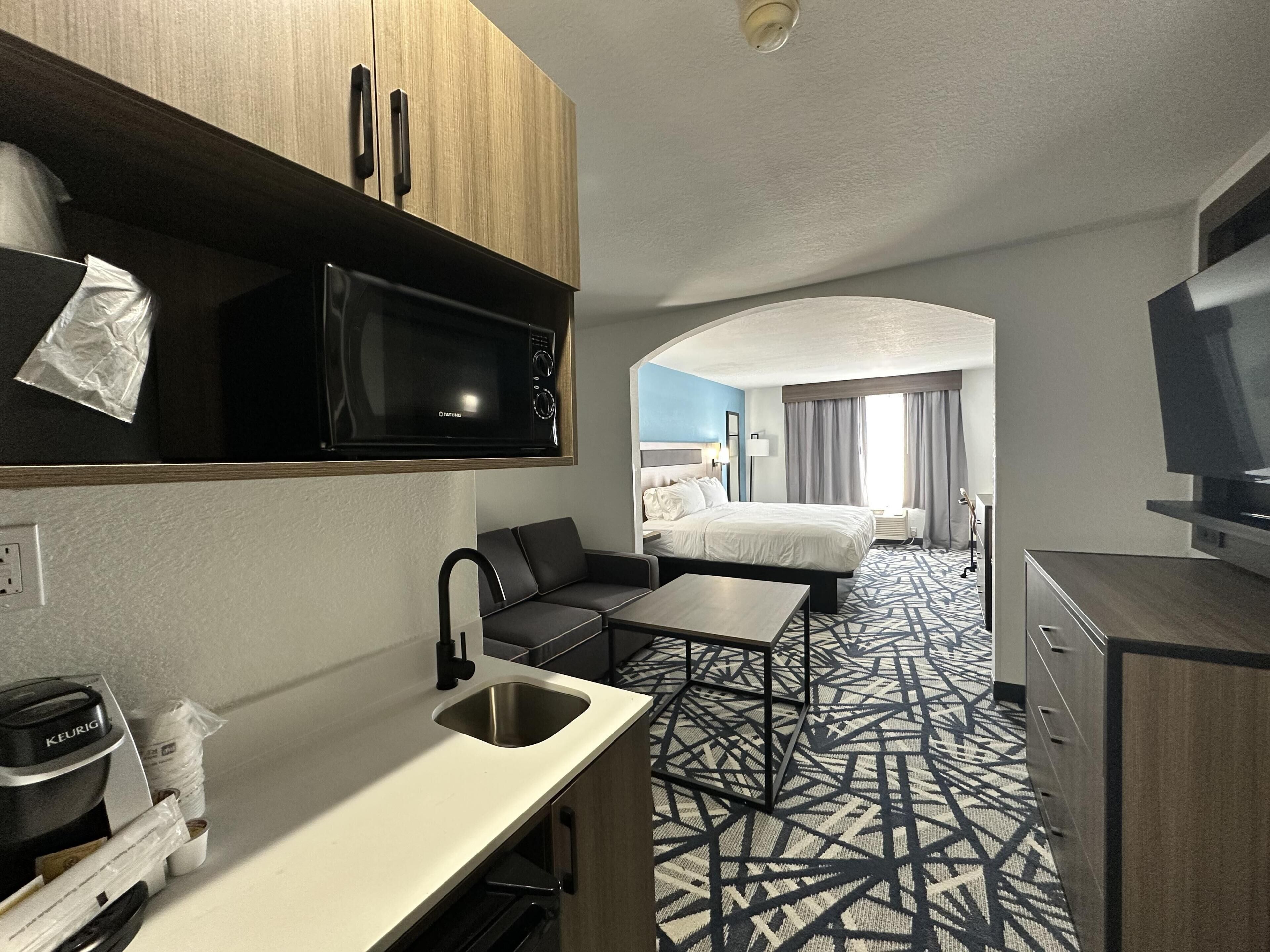 A compact hotel room with a kitchenette, a bed, a TV, and a patterned floor.