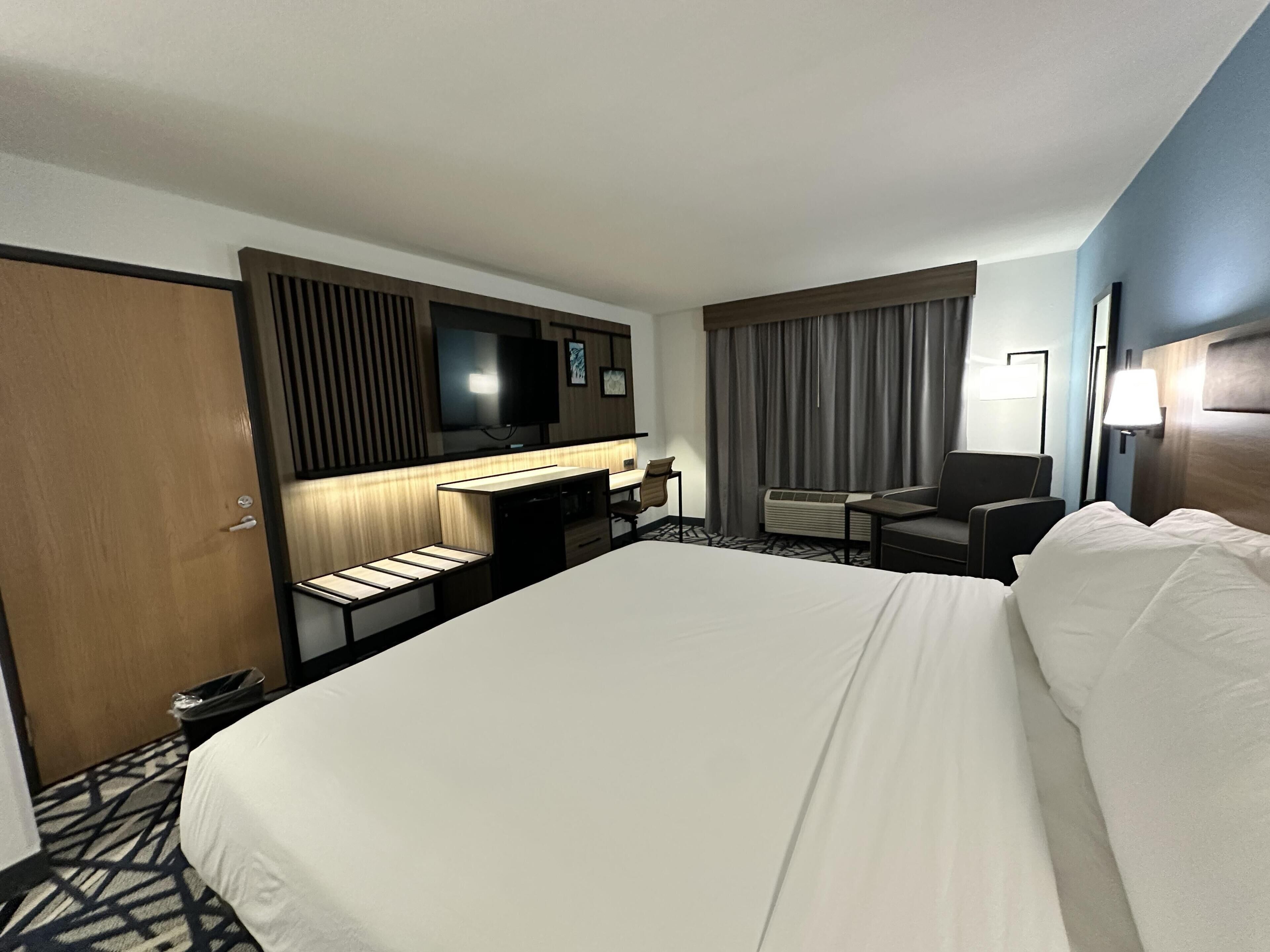 A hotel room with a large bed, a desk with a chair, a television, a window with curtains, and a wall-mounted lamp.