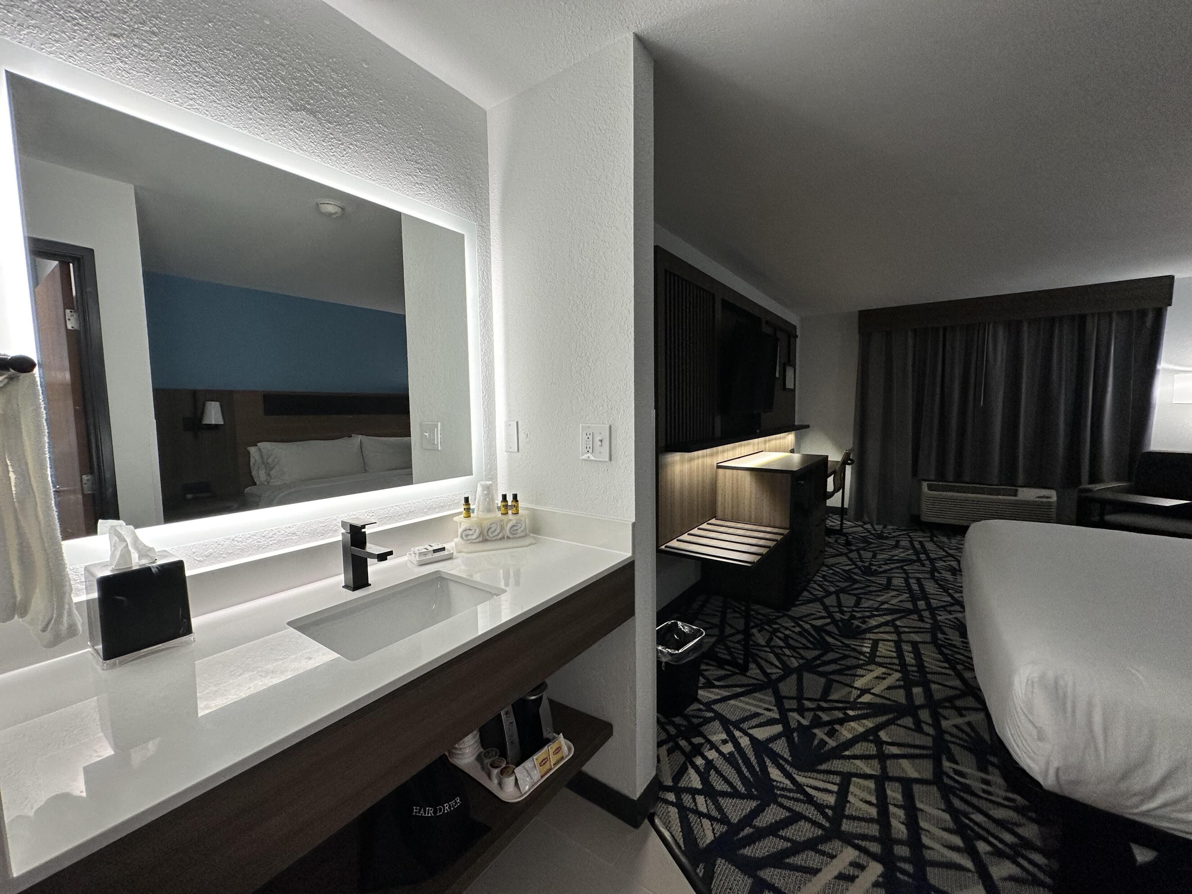 Habitación de hotel con un espejo grande, un lavabo blanco, una cómoda de madera oscura, una cama y una alfombra estampada.