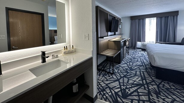 A hotel room with a large mirror, a wooden door, a flat-screen TV, a bed, a desk, and a patterned carpet.