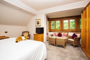 A bedroom with a large bed, a television, wicker furniture, and a view of greenery through the windows.