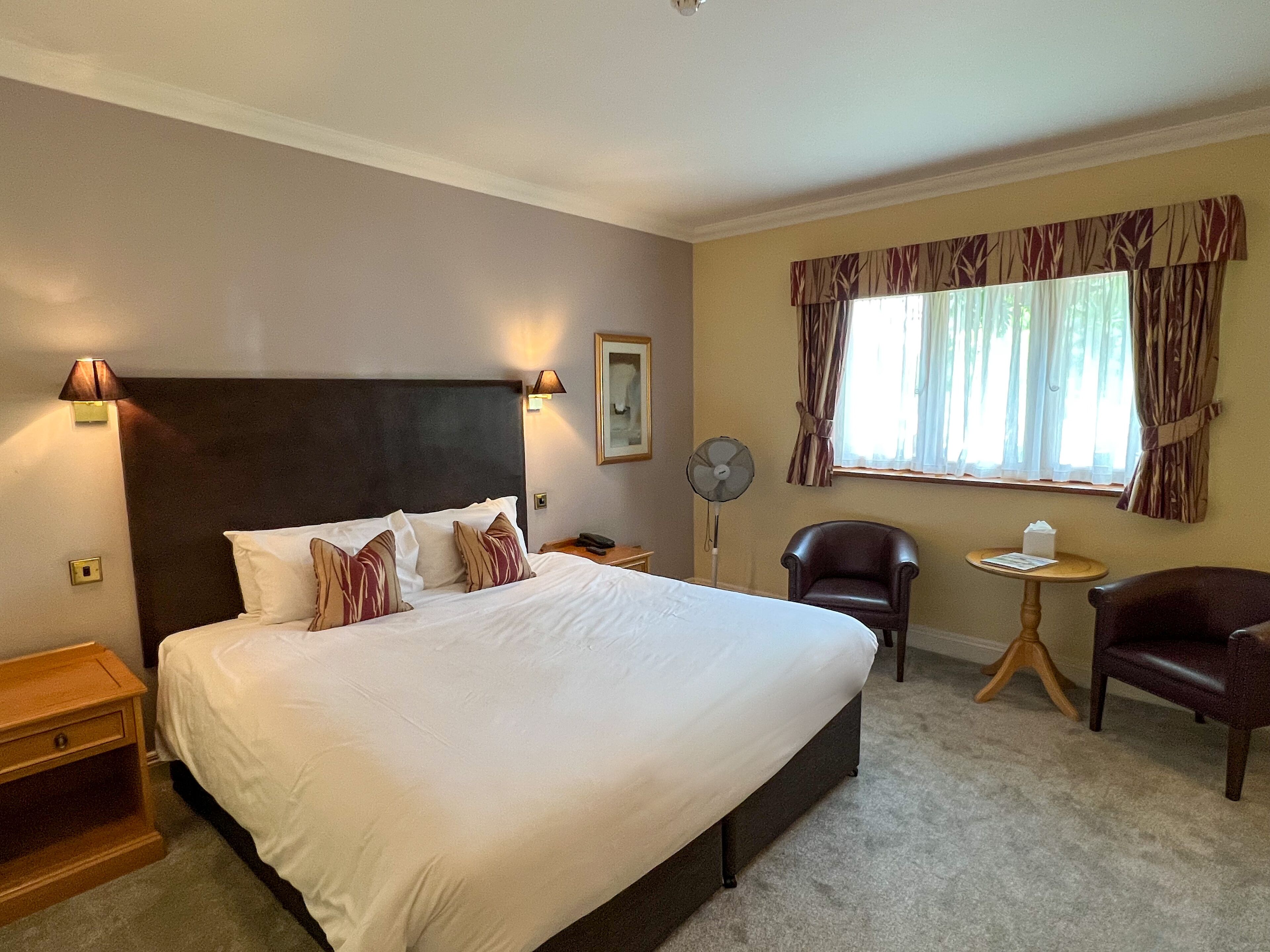 A hotel room with a large bed, two bedside tables, a standing fan, a chair, and a small table with a lamp.